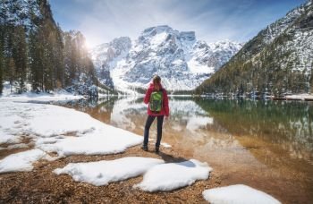 Young woman with backpack on the snowy shore of Braies lake with clear water at sunny bright day in spring. Travel. Landscape with slim girl, reflection in water, mountains, green trees, blue sky