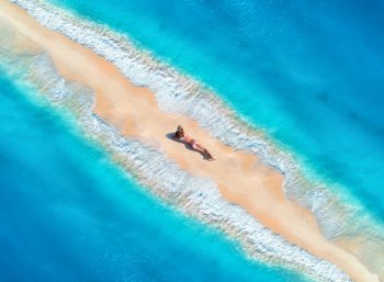 Aerial view of the beautiful young lying woman on the sandy beach and blue sea with waves on the both sides at sunset. Summer holiday. Top view of the back of sporty slim girl, sandbank, clear water. Aerial view of the beautiful young lying woman on the sandy beac