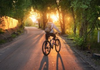 Woman riding a bicycle on the rural road at sunset in summer. Colorful landscape with sporty girl with backpack riding a mountain bike, road, green trees and gold sunlight. Sport and travel. Cycle