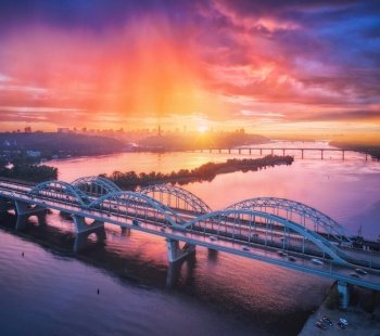Aerial view of beautiful bridge at sunset in Kiev, Ukraine. Landscape with bridge, river, city, colorful sky with red clouds in summer. Cityscape with road, buildings, reflection in water. Top view