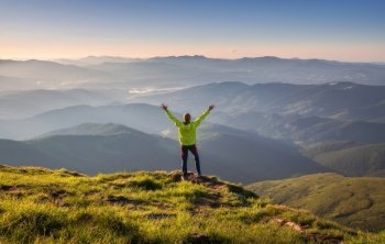 Sporty man standing on the mountain peak with raised up arms against mountain valley in fog at sunset in autumn. Happy young man, rocks, forest and blue sky in fall. Traveler hiking in mountains. Man standing on the mountain peak with raised up arms against