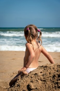 Baby girl in summertime rear view. Girl child relax alone at seaside. Preschool blonde looking at the ocean waves. Adorable little kid sitting back at pile of sand. Pensive child at seashore scenery. 