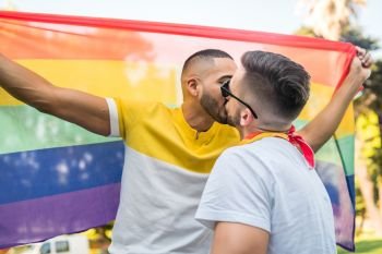 Portrait of young gay couple kissing and showing their love with rainbow flag in the stret. LGBT and love concept.
