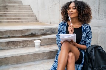Portrait of young afro-american woman thinking while sitting outdoors on stairs with notebook and pen.