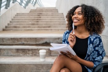 Portrait of young afro-american woman thinking while sitting outdoors on stairs with notebook and pen.