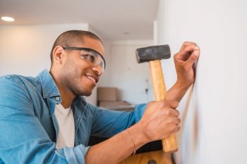 Portrait of young latin man hammering nail on the wall at home. Home improvement and repair home concept.