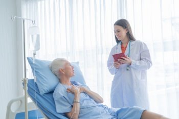 Happy woman doctor talking and using tablet to sick old female senior elderly patient lying in bed in hospital ward room in medical, technology and healthcare treatment concept. Caucasian people.