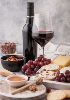 Bottle and glass of red wine with selection of various cheese in wooden box and grapes on light background. Blue Stilton, Red Leicester and Brie Cheese with Cheddar and knife.