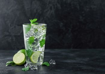 Glass of Mojito cocktail with ice cubes mint and lime on black board with fresh limes. Sparkling refreshing water. Space for text
