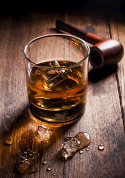 Single malt scotch whiskey in classic glass with ice cubes and smoking pipe on wooden background. Hard light deep shadow