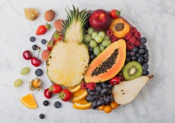 Fresh raw organic summer berries and exotic fruits in white plate on marble background. Pineapple, papaya, grapes, nectarine, orange, apricot, kiwi, pear, lychees, cherry and physalis. Top view