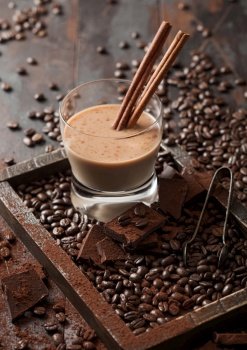 Crystal glass of Irish cream baileys liqueur with cinnamon, coffee beans and powder with dark chocolate in wooden tray on dark wood background. Top view