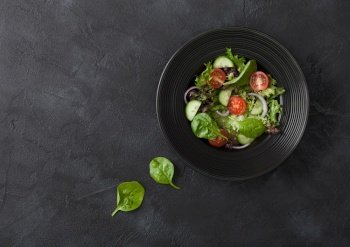 Fresh healthy vegetarian vegetables salad with tomatoes and cucumber, red onion and spinach in black bowl on dark table background. Top view.