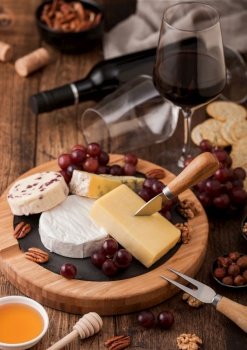 Glass and bottle of red wine with selection of various cheese on the board and grapes on wooden table background. Blue Stilton, Red Leicester and Brie Cheese and knife with linen kitchen cloth.