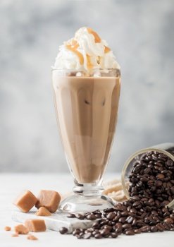 Black iced coffee with fresh milk and caramel with cream in classic milkshake glass and jar of raw coffee beans on light table background.