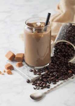 Iced cold coffee with milk and glass container of beans and spoon with salted caramel on marbel board and light table background.