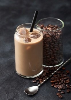 Glass of iced cold coffee and milk with free raw coffee beans in glass container with long spoon on black background.