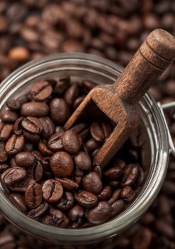 Glass jar with roasted coffee beans and wooden scoop on brown background. Macro