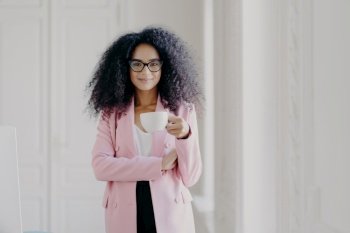 Shot of attractive woman with Afro hairstyle, drinks aromatic beverage, wears glasses, formal wear, poses in office over white background, copy space for your promotional content. People and business