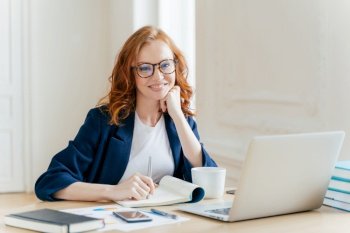 Successful economist in eyewear, checks information on laptop computer, reads information on financial website, writes in notepad and drink hot coffee, sits at workplace. Business and work concept