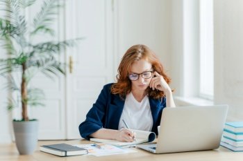 Serious confident female entrepreneur in formal clothes writes down information while watches business webinar, notes main ideas, sits at work place in front of opened computer makes notes for startup