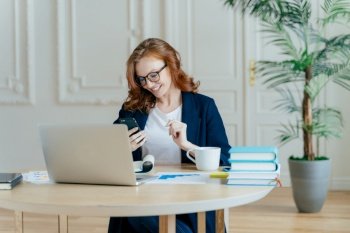 Smiling red haired female focused in modern cellular, happy to recieve text message, poses at workplace, drinks coffee, works on financial project gets ready for video conference with business partner