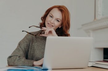Photo of cheerful redhead female student makes research for homework, sits at coworking space in study room, works on laptop computer, uses internet. Freelance web designer creats design for client