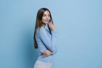 Profile shot of lovely young European woman has slim figure, dressed in blue jumper and white trousers, models in studio during photoshoot, has well groomed face, healthy skin, wears makeup.
