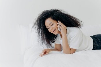 Pleased relaxed woman with curly hair has pleasant telephone conversation, speaks with friend during weekend before sleep, wears white t shirt, lies in cozy bed, keeps modern gadget near ear