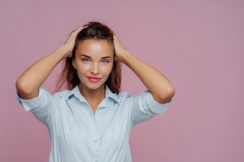 Isolated shot of attractive young woman keeps both hands on hair, looks pleasantly at camera, has natural beauty, wears light blue shirt, poses against purple background, empty space for your text