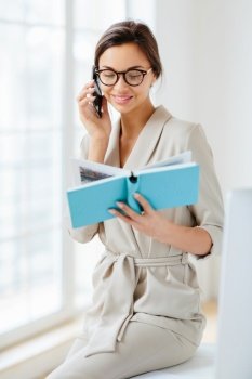 Vertical shot of female administrative manager involved in working process, holds notepad with written notes, discusses something via cellphone, wears spectacles and elegant costume, smiles gently