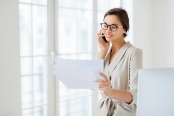 Positive woman in formal clothes, calls to business partner, discusses financial startup project, reads information from papers, wears optical glasses, stands in office, works at international company