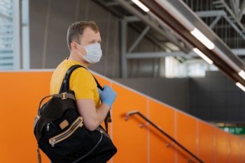 People, health care and coronavirus outbreak concept. Young man wears medical mask and gloves to prevent from pneumonia outbreak, carries backpack on shoulder, concentrated aside, going to travel