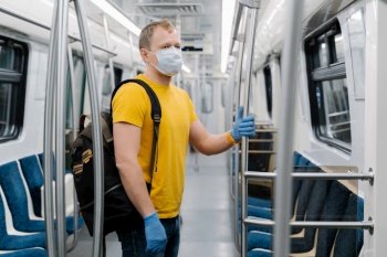 Shot of man wears disposable medical mask during coronavirus outbreak, keeps safety, poses in empty carriage of underground, carries rucksack, commutes to work. Covid-19 and health care concept