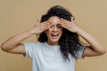 Horizontal shot of happy dark skinned woman covers eyes and smiles, has fun and hides face, has curly hair, dressed in casual white t shirt, isolated on brown background, waits for surprise.