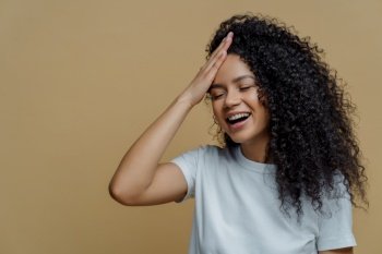 Horizontal shot of happy positive Afro American woman has fun, keeps hand on forehead, giggles happily, closes eyes, has bushy curly hair, wears casual white t shirt, isolated on beige background.