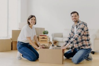 Positive happy husband and wife stand on knees near carton box with cute dog, enjoy relocation in new apartment, smiles pleasantly at camera. Pedigree pet rides in cardboard container on floor