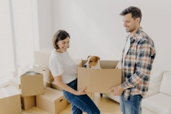 Indoor shot of positive female and male carry carton box with favourite pet, relocate in other place of living, busy with unpacking personal belongings, pose in empty room with many containers.