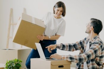 Happy young man points into laptop computer, searches new design for flat, busy woman carries boxes, makes purchase on new habitation, search home decorations, connected to wirless internet.