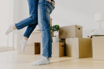 Faceless young couple move in new home, man lifts woman, have fun, surrounded with unpacked cardboard boxes, start new life in recently bought abode. Family, relocation and moving day concept