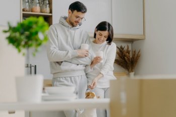 Horizontal view of happy young woman and man play with dog, drink takeaway coffee, stand against kitchen interior, unpack boxes with personal stuff, wear casual clothes, bought new apartment