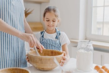Curious small female helper looks attentively how mother cooks, helps to whisk ingredients, wears white t shirt and striped apron, prepare tasty cake together, like baking something delicious
