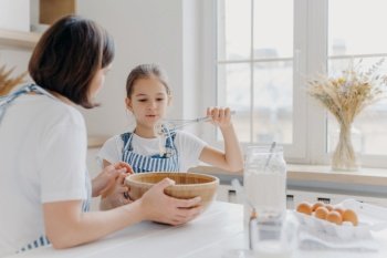 Adorable little child in apron shows whisk with white cream, wears apron, cooks together with mommy, pose in spacious kitchen together, make cake for special event. Children, help about house