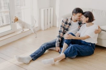 Horizontal shot of pleased husband and wife embrace and kiss, have good relationship, sit on wooden floor near couch, enjoy being at home during weekend, live in new apartment, start new life in abode
