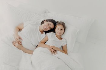 Top view of happy mother and daughter awake in good mood, feel relaxed, enjoy comfort in bed, rejoice good morning, dressed in pyjamas, lie under white blanket on soft pillows. Bedding time.