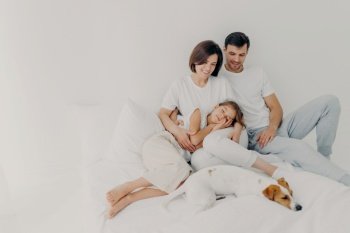 Photo of lovely family spend free time together, stay in bed for long time, had healthy sleep, pedigree dog lies near owners, enjoy comfort and relaxation, blank space on light background for promo