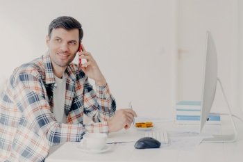 Horizontal shot of busy male freelancer has telephone conversation, dressed in checkered shirt, writes down information, sits at desktop with computer, solves financial problem, poses at workplace