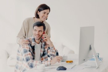 Happy young man holds mobile phone, discusses issues, manages home accounts with wife, pose in coworking space, sit in front of computer, discuss family expenses. Couple collaborate together