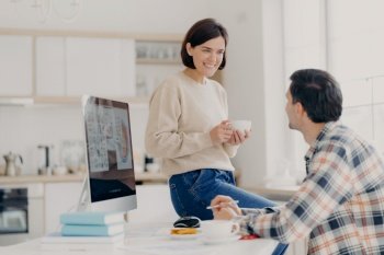 Positive young woman and man discuss renting apartment, pose in coworking space with computer and paper documents, happy wife drinks coffee, looks with pleasant smile at husband, manage budget