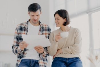 Serious couple study documents together, have serious looks, drink coffee, dressed in casual wear, plan their budget, pose in spacious light room, do paperwork, busy preparing financial report